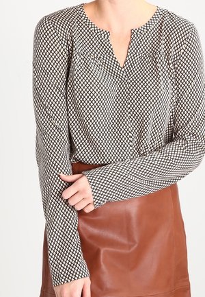 Person wearing a long-sleeve patterned blouse tucked into a brown leather skirt, with one arm crossed and hand gently touching the opposite sleeve.