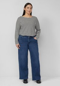 Bluzka w czarno-białe paski z długim rękawem, zestawiona z szerokimi, ciemnoniebieskimi dżinsami. Model stoi z rękami w kieszeniach, ma na nogach czarne buty.