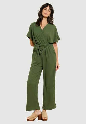 Vrouw draagt olijfgroene wikkel-jumpsuit met wijde pijpen en een gestrikte tailleceintuur, gecombineerd met beige hoge hakken open-toe klompen.