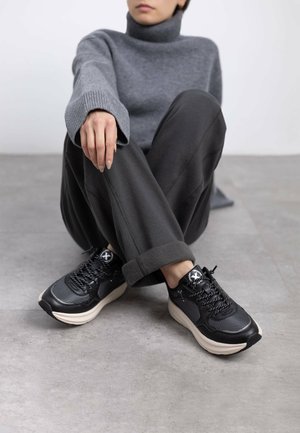 Zapatillas negras con malla y cuero, con una suela gruesa color crema, forma redondeada y cordones. Se llevan con un suéter de cuello alto gris y pantalones oscuros.