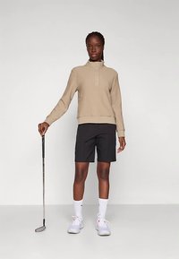 Sudadera beige texturizada con cuello y tres botones, combinada con pantalones cortos negros. Persona sostiene un palo de golf, lleva zapatillas deportivas blancas.