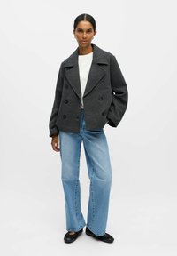 Veste grise à double boutonnage avec larges revers, boutons noirs, assortie à une chemise blanche et un jean à larges jambes bleu clair. Chaussures plates noires.