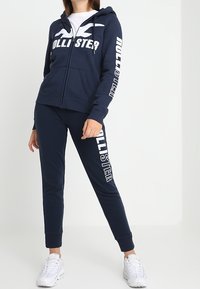 Marinblå zip-up hoodie och matchande joggers, båda med "Hollister"-logotyp i vitt. Materialet är mjukt, med en avslappnad passform, huva och fickor.
