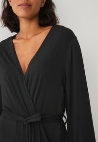 Robe noire portefeuille en tissu doux, avec un décolleté en V profond, des manches longues et une taille nouée pour un ajustement réglable. Design simple et minimaliste.