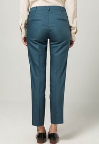 Teal op maat gemaakte broeken met een slim fit, een gladde textuur en achterzakken, gecombineerd met donkere schoenen en een lichtgekleurde blouse.