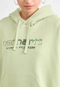 Lys grønn hettegenser med kengurulomme, hettekrage og blank präget "carhartt" tekst. Mykt stoff med en glatt tekstur.