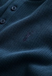 Pulover tricotat de culoare albastru marin, cu un material texturat, două butoane negre și un logo discret embosat aproape de guler.