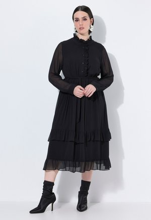 Vestido negro de manga larga con acentos de volantes, cierre de botones en la parte delantera, cintura ajustada y falda en capas. Combinado con botas negras hasta el tobillo.