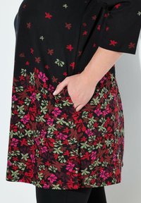 Zwarte tuniek met een bloemenpatroon in rood, roze en groen, met een zak aan de rechterkant en een relaxte pasvorm.