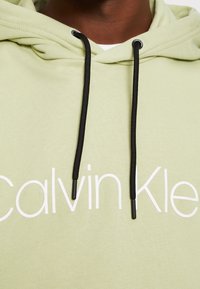 Ljusgrön hoodie med svarta dragsnören. Har framträdande vit text som säger "Calvin Klein" på framsidan. Mjuk tygtextur.