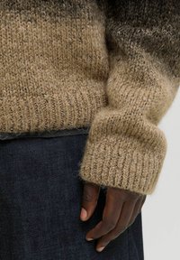 Nahaufnahme einer dunkelhäutigen Hand neben dem Ärmel eines beige-braunen Strickpullovers und dunklen Denim-Stoff.