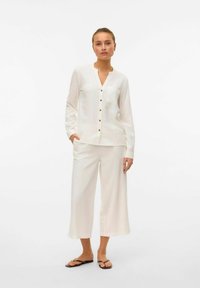 Camicia bianca con colletto a V e taschino, abbinata a pantaloni bianchi a gamba larga e lunghezza alla caviglia. Modello con sandali neri.