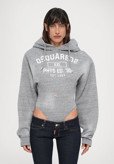 Persoon in een grijze versleten hoodie bodysuit met witte tekst, gecombineerd met donkerblauwe jeans, staande tegen een witte achtergrond.