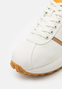 Λευκά αθλητικά παπούτσια με λείο δέρμα στην επάνω επιφάνεια, που διαθέτουν μπεζ λωρίδα, στρογγυλή μύτη και πορτοκαλί εσωτερικό. Η εξωτερική σόλα από καουτσούκ έχει υφή με κυκλικά μοτίβα.