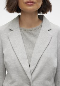 Blazer gris claro con solapas de pico, cierre de botón y textura suave, superpuesto a una blusa gris de canalé. Diseño simple y entallado.