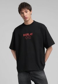 T-shirt nera oversize in cotone, con testo e simboli rossi "REPLAY" sul davanti. Maniche corte e scollatura rotonda.