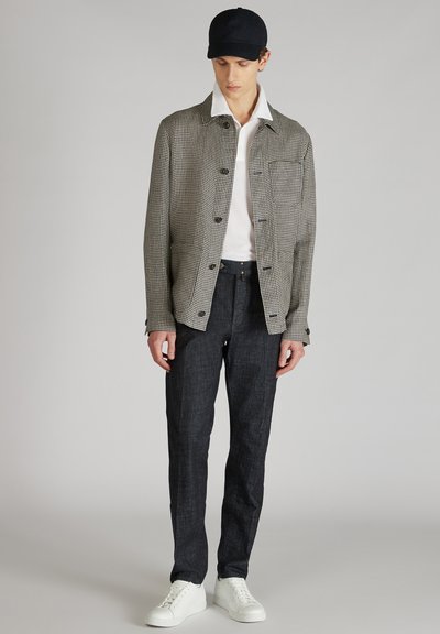 Hondstooth-gepatroneerde jas, witte kraag, wit T-shirt, donkere denim broek en witte sneakers, gedragen door een model tegen een effen achtergrond.