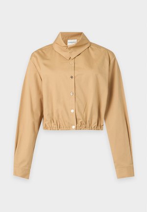Vrouw draagt een beige cropped jas, een wit gestructureerd topje, donkerblauwe knielange shorts, zwarte loafers en draagt een bordeauxrode handtas op haar schouder.