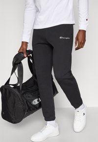 Μαύρο παντελόνι φόρμας από φλίς με ελαστικά μανσέτες και λευκό λογότυπο "Champion", σε συνδυασμό με μαύρη τσάντα duffle και λευκά αθλητικά παπούτσια.