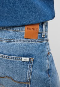Blå denimjeans har en brun läderetikett med "MUSTANG" präglat, kontrasterande sömmar och en liten logotagg på bakfickan.