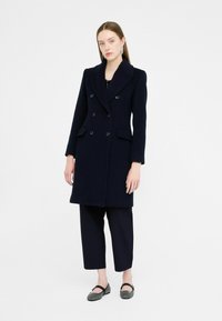 Marineblauwe wollen dubbelzijdige jas met notch-lapels, twee voorzakken en knoopsluiting, gecombineerd met zwarte cropped broek en platte schoenen.