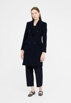 Cappotto doppiopetto in lana blu navy con revers, due tasche frontali e chiusura con bottoni, abbinato a pantaloni neri cropped e scarpe basse.
