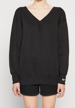 Persoon draagt een zwart sweatshirt met lange mouwen en V-hals en zwarte shorts, met een wit Nike-logo op de linkermouwmanchet.