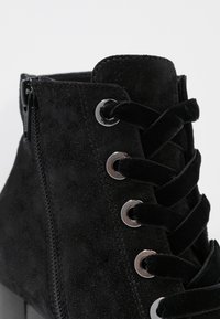 Bottine en suède noir avec un col haut, des œillets en métal et des lacets plats, dotée d'une fermeture éclair sur le côté pour un enfilage facile. Détail de surface texturée.