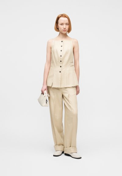 Γυναίκα που φοράει μπεζ αμάνικο τοπ με κουμπιά και αντίστοιχο φαρδύ παντελόνι, κρατώντας μια μικρή μπεζ τσάντα, στέκεται σε λευκό φόντο.