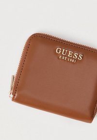 Portafoglio in pelle marrone con cerniera e logo dorati "GUESS EST 1981" sulla parte anteriore. Texture liscia con forma rettangolare.