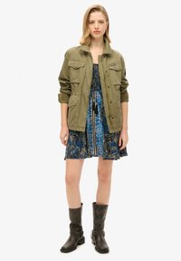 Veste militaire vert olive avec plusieurs poches, portée sur une robe bleue à motif paisley. Modèle portant des bottes en cuir noir usé.