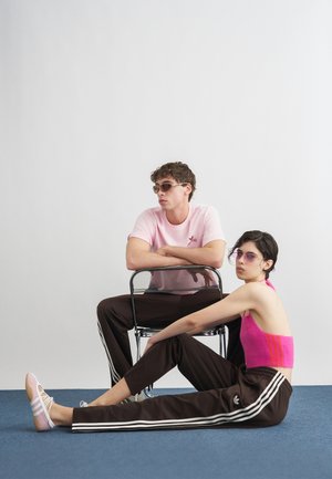 Δύο νέοι ενήλικες με γυαλιά ηλίου και παντελόνια Adidas. Ο ένας κάθεται σε μια καρέκλα με ροζ πουκάμισο, ενώ ο άλλος γέρνει προς τα πίσω με ροζ τοπ πάνω σε μπλε πάτωμα.