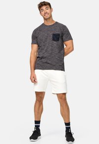 Camiseta de manga corta gris oscuro con un bolsillo a rayas en azul marino, combinada con pantalones cortos blancos. El modelo lleva zapatillas negras y calcetines a rayas en azul marino y blanco.