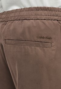 Καφέ παντελόνια με ελαστική μέση, πίσω τσέπη με σχισμή και κεντημένο λογότυπο "Calvin Klein" κοντά στην τσέπη.