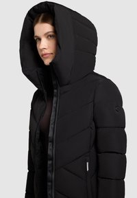 Černá péřová bunda s velkou kapucí, prošívaným designem a zapínáním na zip. Obsahuje kulatou knoflíkovou dekoraci na rukávu a logo štítek.