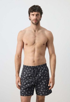 Hombre sin camiseta con cabello corto y oscuro, que lleva puestos pantalones cortos de baño negros con un patrón paisley blanco, de pie frente a un fondo claro y liso.