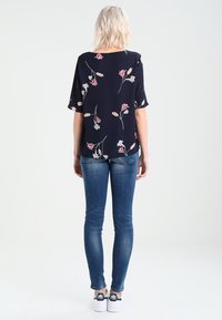 Blusa floreale navy con maniche corte, caratterizzata da motivi floreali rosa e beige, abbinata a jeans skinny blu e sneaker bianche.