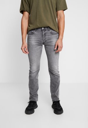 Jeans gris ajustés avec un délavage, dotés de cinq poches et d'une fermeture à bouton. Associés à un t-shirt vert olive et des baskets noires.