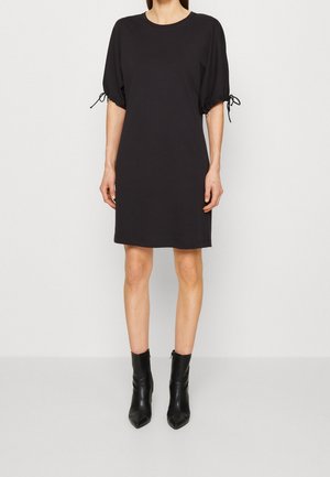 Femme portant une robe noire jusqu'au genou avec des manches mi-longues bouffantes nouées aux extrémités, associée à des bottines noires à bout pointu.