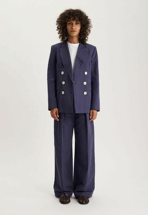 Femme aux cheveux bouclés portant un blazer croisé à rayures bleu marine et un pantalon large avec des chaussures noires sur un fond uni.