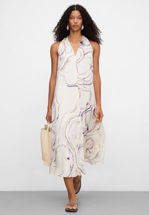 Vrouw met krullend haar, gekleed in een mouwloze witte midijurk met een abstracte print, die een beige totebag vasthoudt en zwarte sandalen draagt.