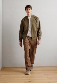 Green suede bomber jacket over a white t-shirt, paired with brown trousers and beige sneakers, set against a neutral background.