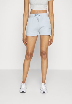 Mujer con pantalones cortos azul claro con cordón, zapatillas blancas y una camiseta sin mangas gris corta, de pie frente a un fondo liso.