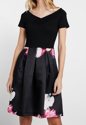 Femme portant une robe noire à manches courtes avec un décolleté épaules dénudées et une jupe plissée ornée de grands motifs floraux roses et blancs.