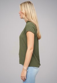 Chemise olive à manches courtes avec un col rond et un tissu texturé. Portée avec un jean bleu clair, vue de côté.