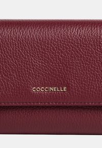 Bordó kožená peněženka s texturovaným povrchem a klopovým uzávěrem. Obsahuje zlaté logo "Coccinelle" a prošívané detaily.