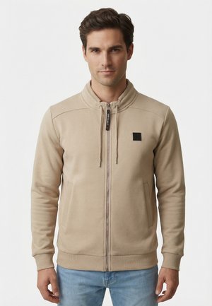 Jeune homme portant un sweat-shirt beige à fermeture éclair avec cordons et un jean bleu clair, debout devant un fond blanc uni.