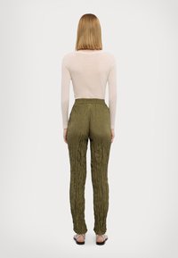 Vrouw met steil blond haar staat met haar rug naar ons toe en draagt een doorschijnende witte top met lange mouwen, olijfgroene gekreukte broek en platte sandalen.