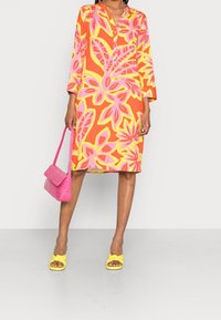 Buntes Kleid mit orange-, pink- und gelbblumigem Muster, 3/4-Arm, V-Ausschnitt, kombiniert mit einer pinken Tasche und gelben Sandalen mit Knoten.