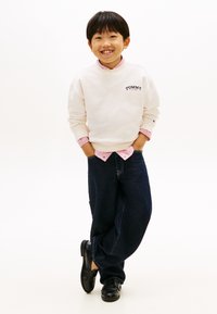 Ανοιχτό μπεζ φούτερ με λογότυπο "TOMMY HILFIGER", από πάνω από κόκκινο και λευκό ριγέ πουκάμισο, συνδυασμένο με σκούρο μπλε τζιν και μαύρα παπούτσια.